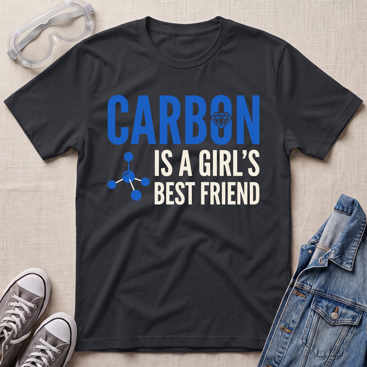 A black graphic t-shirt laid flat on a textured beige fabric background, styled with safety goggles, gray sneakers, and a denim jacket. The t-shirt features the text 'CARBON IS A GIRL'S BEST FRIEND' in blue and white, with a blue molecular structure graphic.
