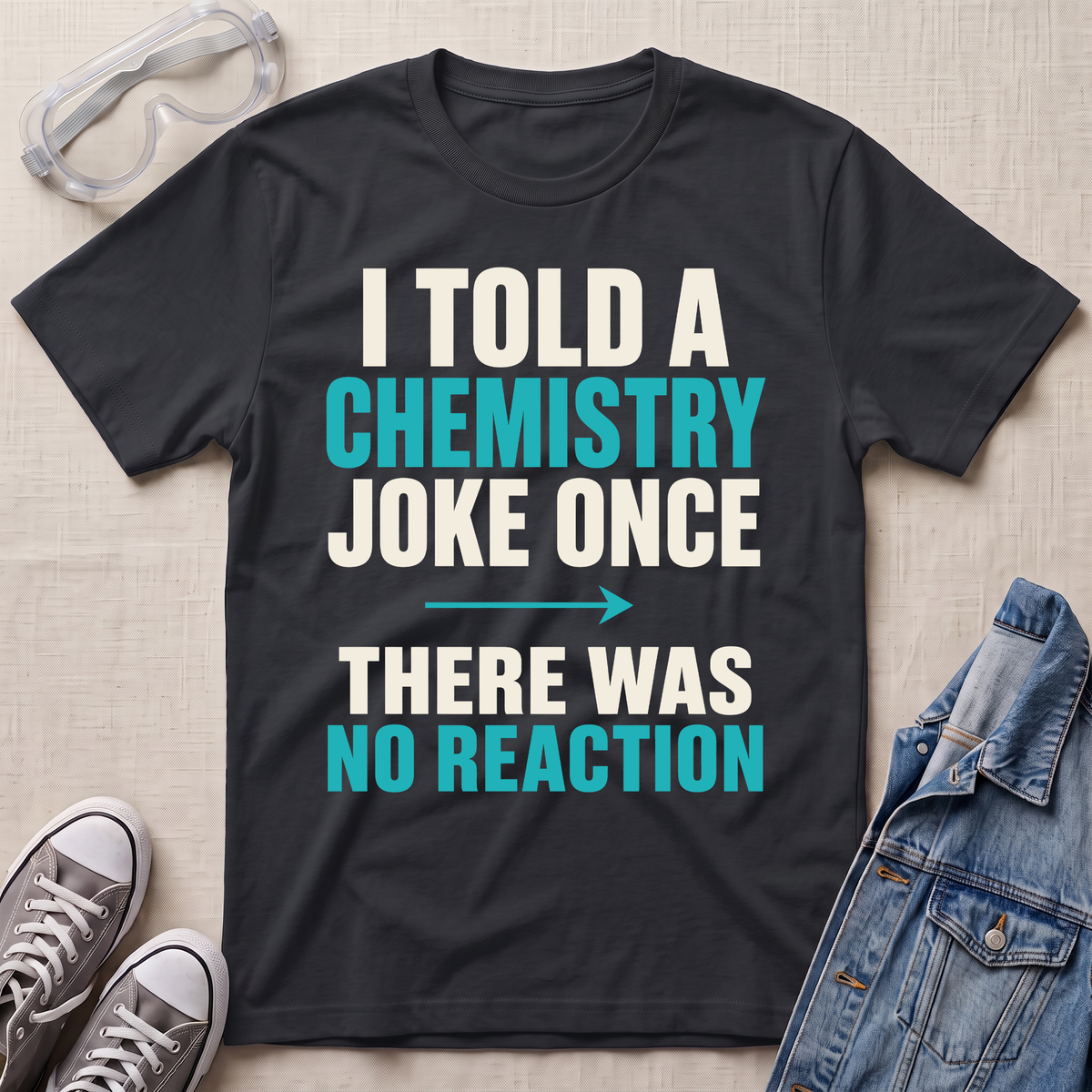 A black graphic t-shirt laid flat on a light beige textured background, surrounded by styling props including safety goggles, gray sneakers, and a denim jacket. The t-shirt features the text 'I TOLD A CHEMISTRY JOKE ONCE THERE WAS NO REACTION' in white and bright teal lettering.