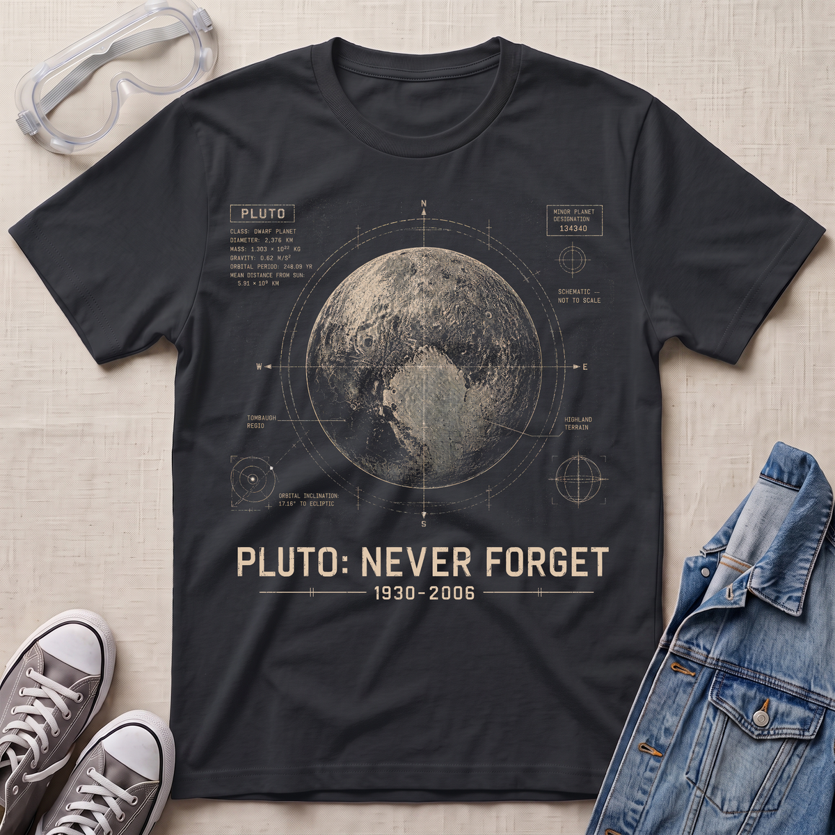 A black graphic t-shirt featuring a vintage-style space blueprint design of Pluto with the text 'PLUTO: NEVER FORGET 1930-2006'. The t-shirt is styled in a flat-lay arrangement on a textured beige fabric background, accompanied by clear safety goggles, gray sneakers, and a blue denim jacket.