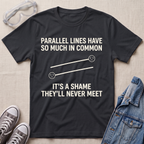 A black graphic t-shirt laid flat on a beige textured background, surrounded by styling props including safety goggles, gray sneakers, and a denim jacket. The t-shirt features a white graphic of two parallel lines with sad faces and the text 'PARALLEL LINES HAVE SO MUCH IN COMMON IT'S A SHAME THEY'LL NEVER MEET'.