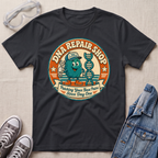 A black graphic t-shirt laid flat on a light beige textured background, surrounded by lifestyle props including safety goggles, gray sneakers, and a denim jacket. The t-shirt features a circular retro-style graphic of a teal cartoon DNA molecule wearing a cap and holding a wrench, with the text 'DNA REPAIR SHOP' and 'Patching Your Base Pairs Since Day One'.