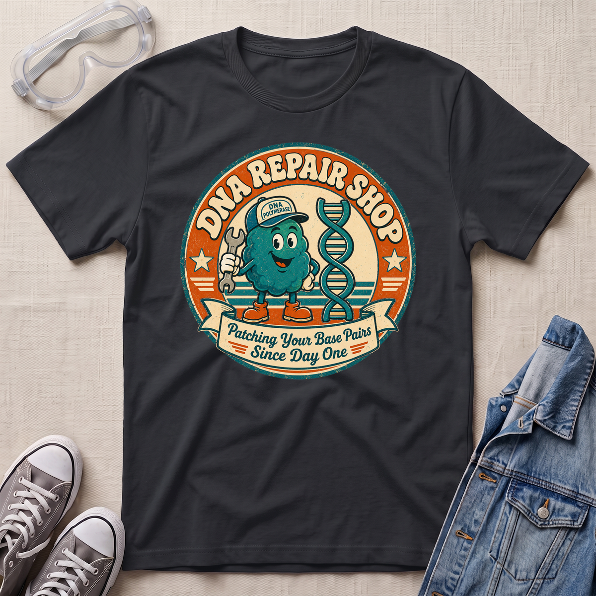 A black graphic t-shirt laid flat on a light beige textured background, surrounded by lifestyle props including safety goggles, gray sneakers, and a denim jacket. The t-shirt features a circular retro-style graphic of a teal cartoon DNA molecule wearing a cap and holding a wrench, with the text 'DNA REPAIR SHOP' and 'Patching Your Base Pairs Since Day One'.