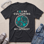 A black graphic t-shirt laid flat on a light beige textured background, surrounded by styling props including safety goggles, gray sneakers, and a denim jacket. The t-shirt features a circular petri-dish graphic filled with teal microorganisms and white text that reads 'YOU'RE CULTURING MY INTEREST'.