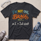 A black graphic t-shirt laid flat on a light beige textured background, surrounded by styling props including clear safety goggles, gray sneakers, and a blue denim jacket. The t-shirt features a colorful text graphic reading 'I'M NOT ONE TO BRAGG BUT.