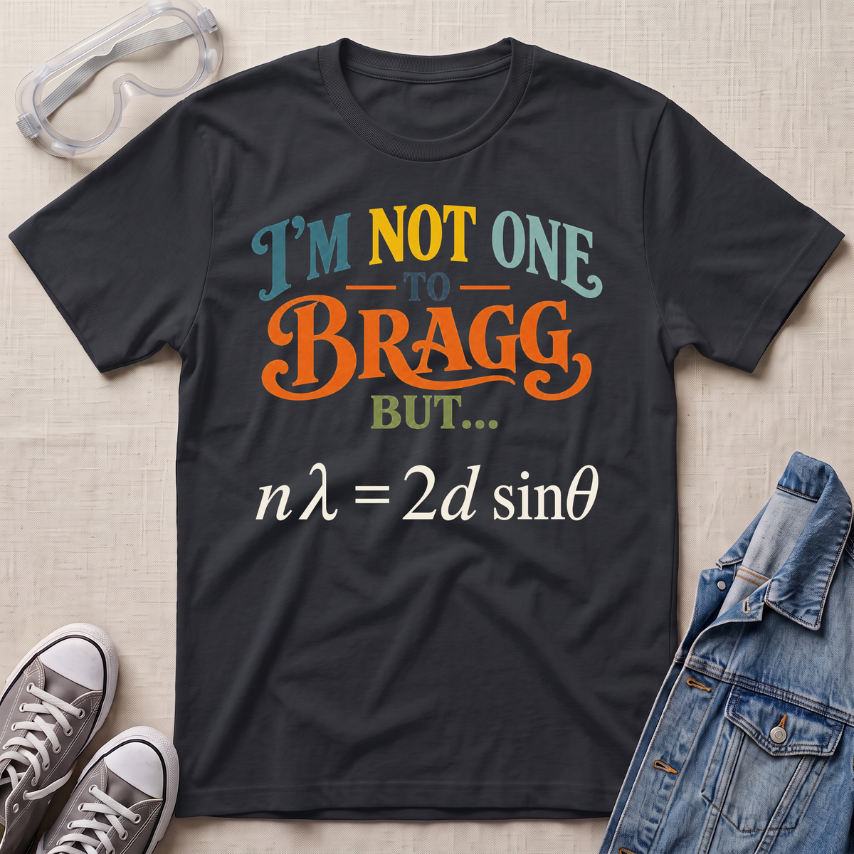 A black graphic t-shirt laid flat on a light beige textured background, surrounded by styling props including clear safety goggles, gray sneakers, and a blue denim jacket. The t-shirt features a colorful text graphic reading 'I'M NOT ONE TO BRAGG BUT.