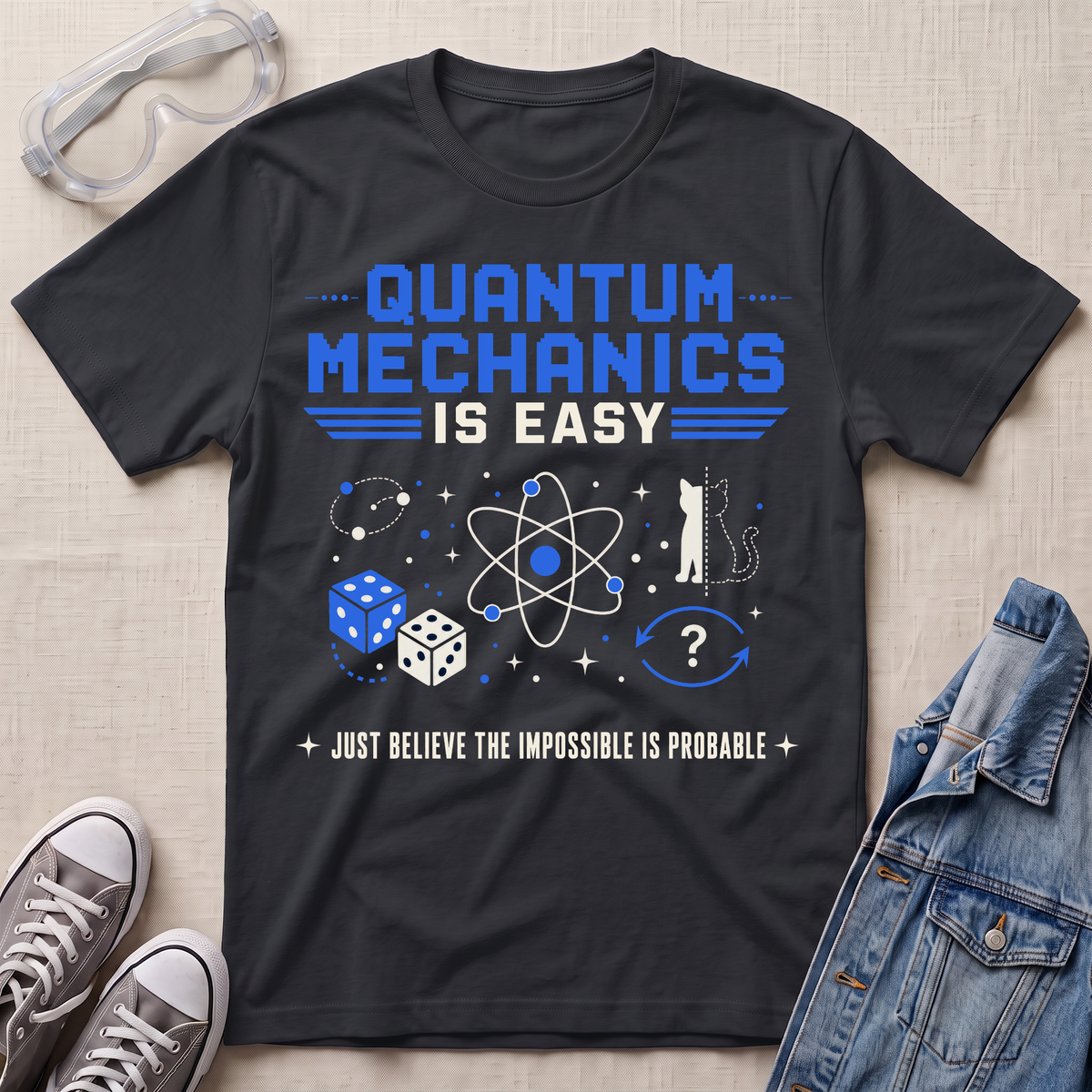 A black graphic t-shirt laid flat on a light beige textured background, styled with safety goggles, gray sneakers, and a denim jacket. The t-shirt features a blue and white graphic that reads 'QUANTUM MECHANICS IS EASY' with scientific doodles and the phrase 'JUST BELIEVE THE IMPOSSIBLE IS PROBABLE'.