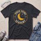 A black t-shirt with a yellow and cream graphic reading 'SPACE CHIMP ACADEMY' around a crescent moon and stars. The shirt is styled in a flat lay with a pair of safety goggles, gray sneakers, and a denim jacket on a light beige textured background.