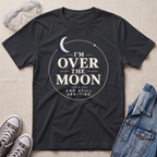 A black graphic t-shirt laid flat on a light beige textured background, surrounded by lifestyle props including clear safety goggles, gray sneakers, and a blue denim jacket. The t-shirt features a circular graphic with a crescent moon and the text 'I'M OVER THE MOON AND STILL ORBITING'.