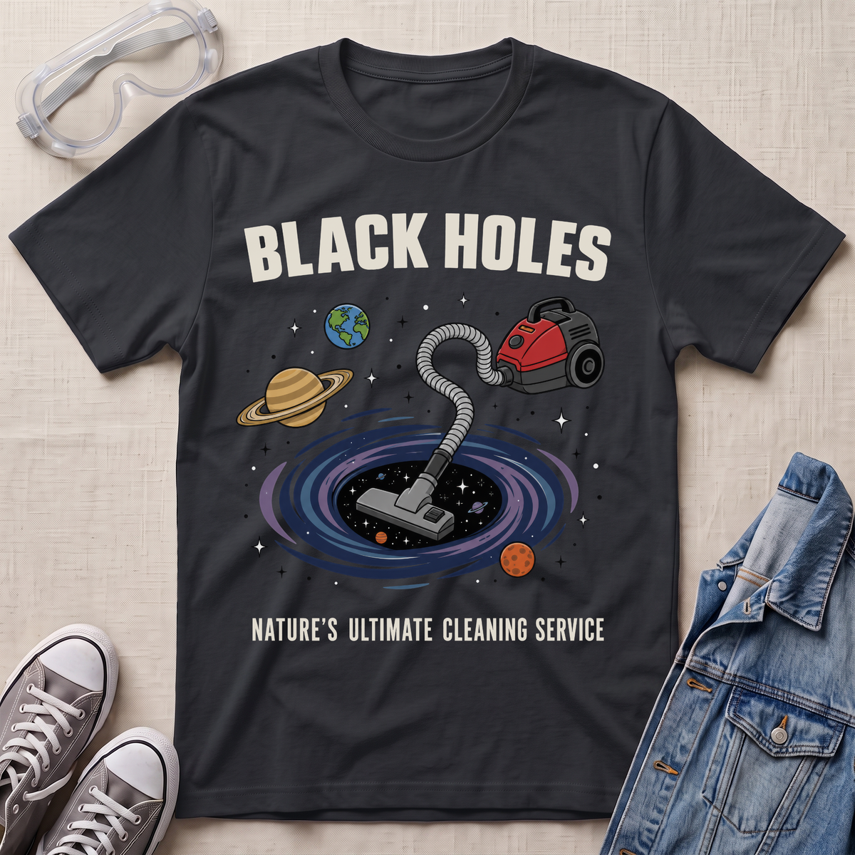 A black graphic t-shirt laid flat on a textured fabric background, styled with safety goggles, sneakers, and a denim jacket. The t-shirt features a cartoon illustration of a vacuum cleaner falling into a black hole surrounded by planets, with the text 'BLACK HOLES' and 'NATURE'S ULTIMATE CLEANING SERVICE'.