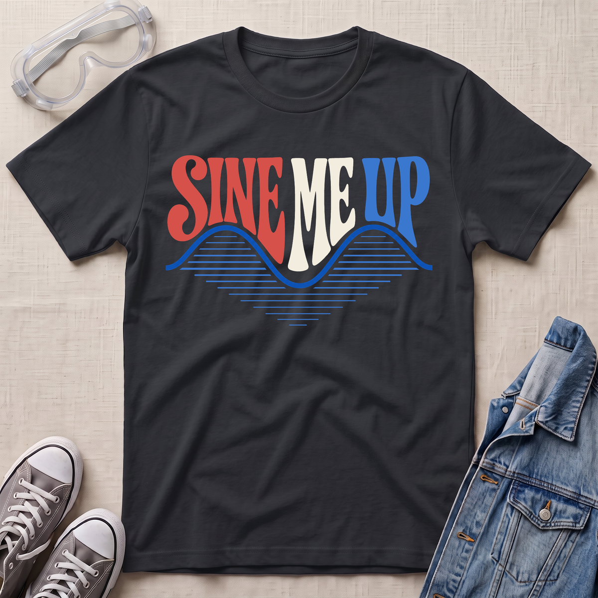 A black graphic t-shirt laid flat on a light beige textured background, styled with safety goggles, gray sneakers, and a denim jacket. The t-shirt features a retro-style graphic reading 'SINE ME UP' in red, white, and blue, with a blue wave design underneath.