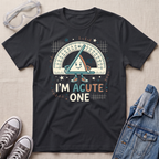 A black graphic t-shirt laid flat on a light beige textured background, surrounded by styled props including safety glasses, gray sneakers, and a blue denim jacket. The t-shirt features a cartoon triangle protractor character holding a ruler, with the text 'I'M ACUTE ONE.