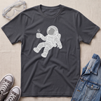A charcoal gray t-shirt featuring a white line-art graphic of an astronaut floating and holding a steaming mug. The t-shirt is styled in a flat-lay arrangement on a light beige textured background, surrounded by props including safety goggles, gray sneakers, and a blue denim jacket.