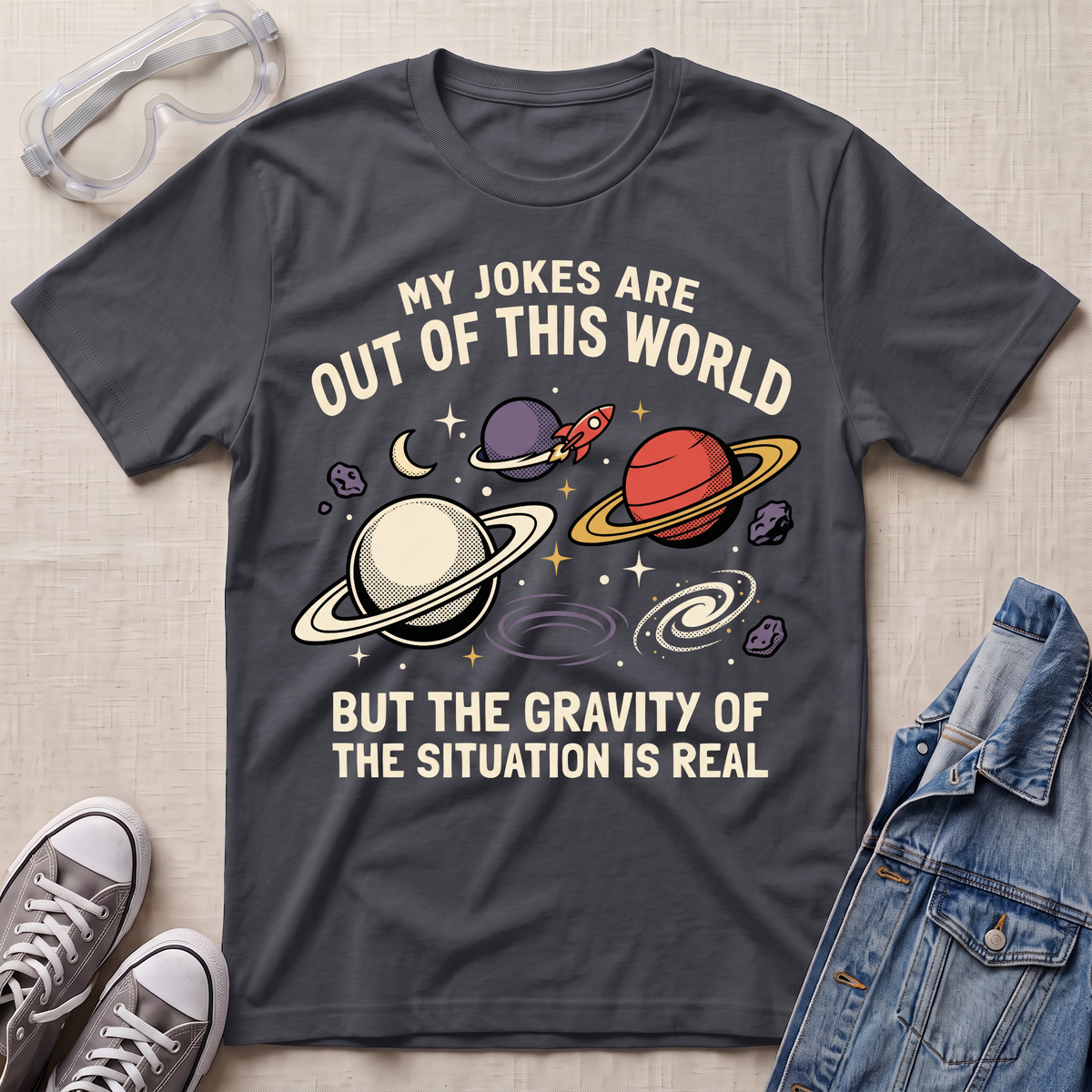 A dark charcoal gray graphic t-shirt laid flat on a light beige textured background, surrounded by styling props including clear safety glasses, gray sneakers, and a blue denim jacket. The t-shirt features a colorful space-themed graphic with planets, stars, and the text 'MY JOKES ARE OUT OF THIS WORLD BUT THE GRAVITY OF THE SITUATION IS REAL'.