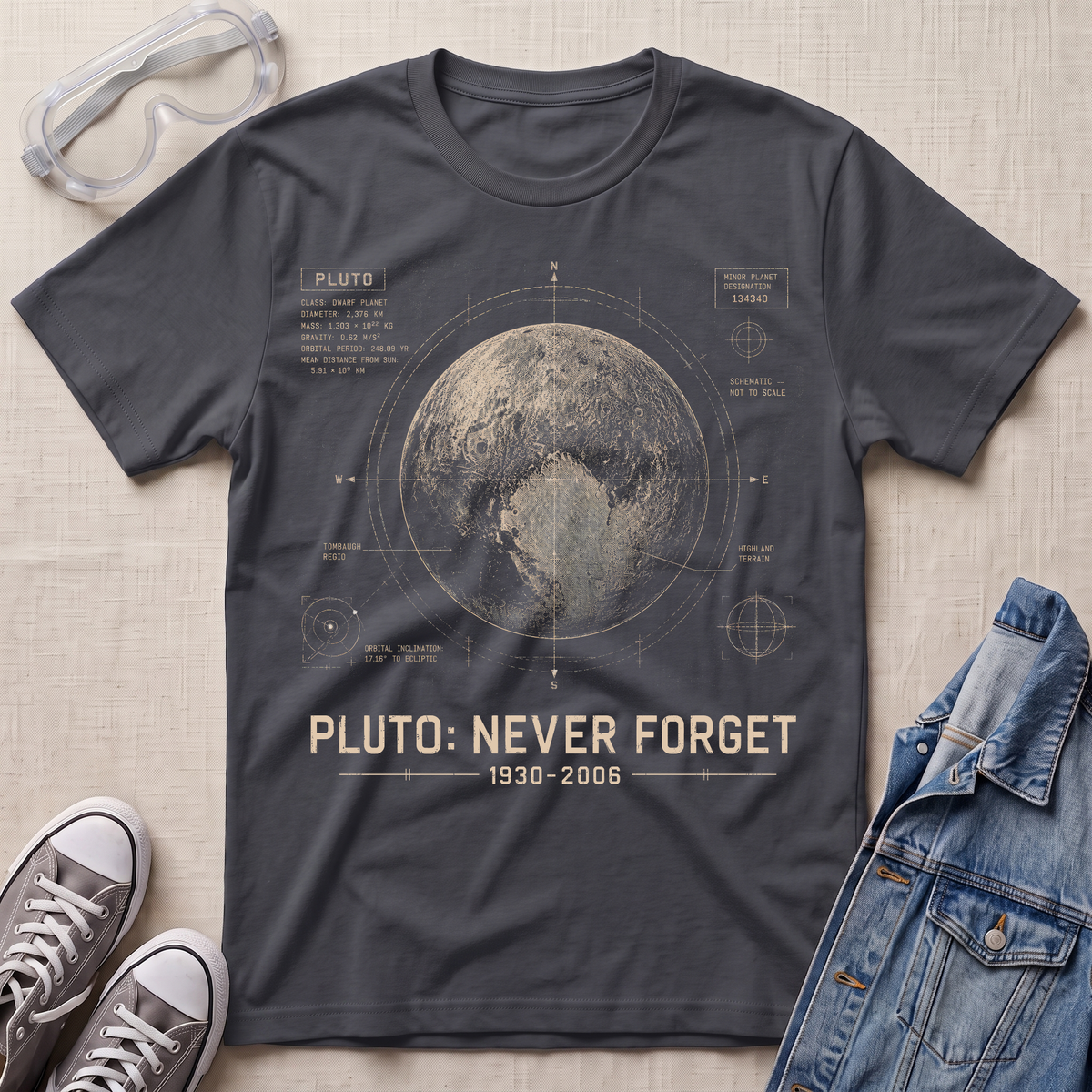 A dark charcoal gray graphic t-shirt featuring a vintage-style design of Pluto with the text 'PLUTO: NEVER FORGET 1930-2006'. The t-shirt is displayed in a flat-lay arrangement on a textured beige background, surrounded by props including clear safety goggles, gray sneakers, and a blue denim jacket.