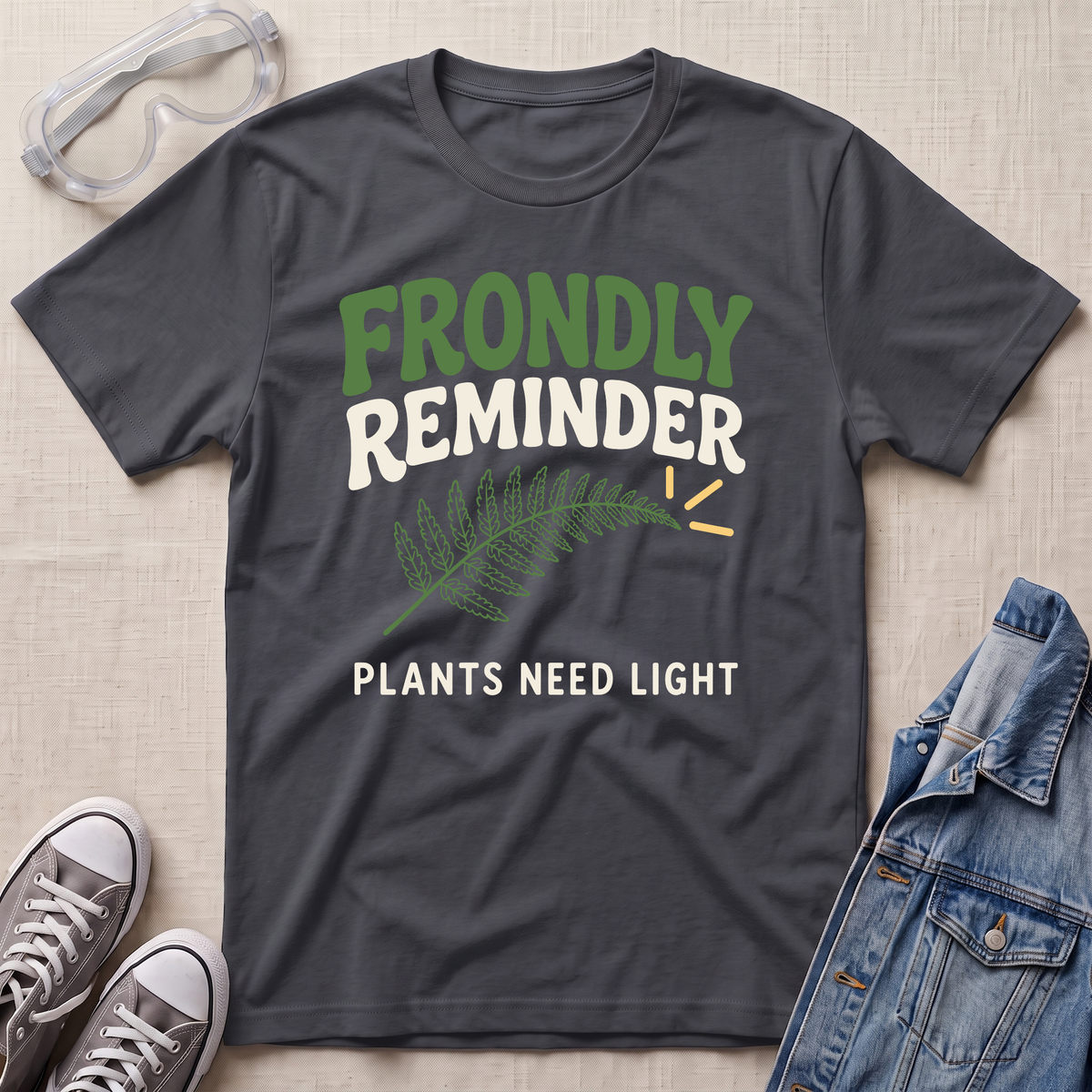 A dark charcoal gray graphic t-shirt laid flat on a light beige textured background, styled with safety goggles, gray sneakers, and a blue denim jacket. The t-shirt features a green fern leaf graphic and the text 'FRONDLY REMINDER PLANTS NEED LIGHT' in green and white.