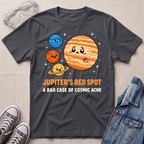 A dark charcoal gray graphic t-shirt laid flat on a light beige textured background, styled with clear safety goggles, gray sneakers, and a blue denim jacket. The t-shirt features a cartoon graphic of Jupiter and other planets with the text 'JUPITER'S RED_spot A BAD CASE OF COSMIC ACNE'.