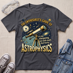 A dark charcoal gray graphic t-shirt laid flat on a light beige textured background, styled with safety goggles, gray sneakers, and a denim jacket. The t-shirt features a detailed space-themed graphic with a telescope, planets, stars, and the text 'THE ENTHUSIAST'S GUIDE TO ASTROPHYSICS' in a bold yellow font.