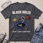 A charcoal graphic t-shirt laid flat on a textured fabric background, styled with safety goggles, sneakers, and a denim jacket. The t-shirt features a cartoon illustration of a vacuum cleaner falling into a black hole surrounded by planets, with the text 'BLACK HOLES' and 'NATURE'S ULTIMATE CLEANING SERVICE'.