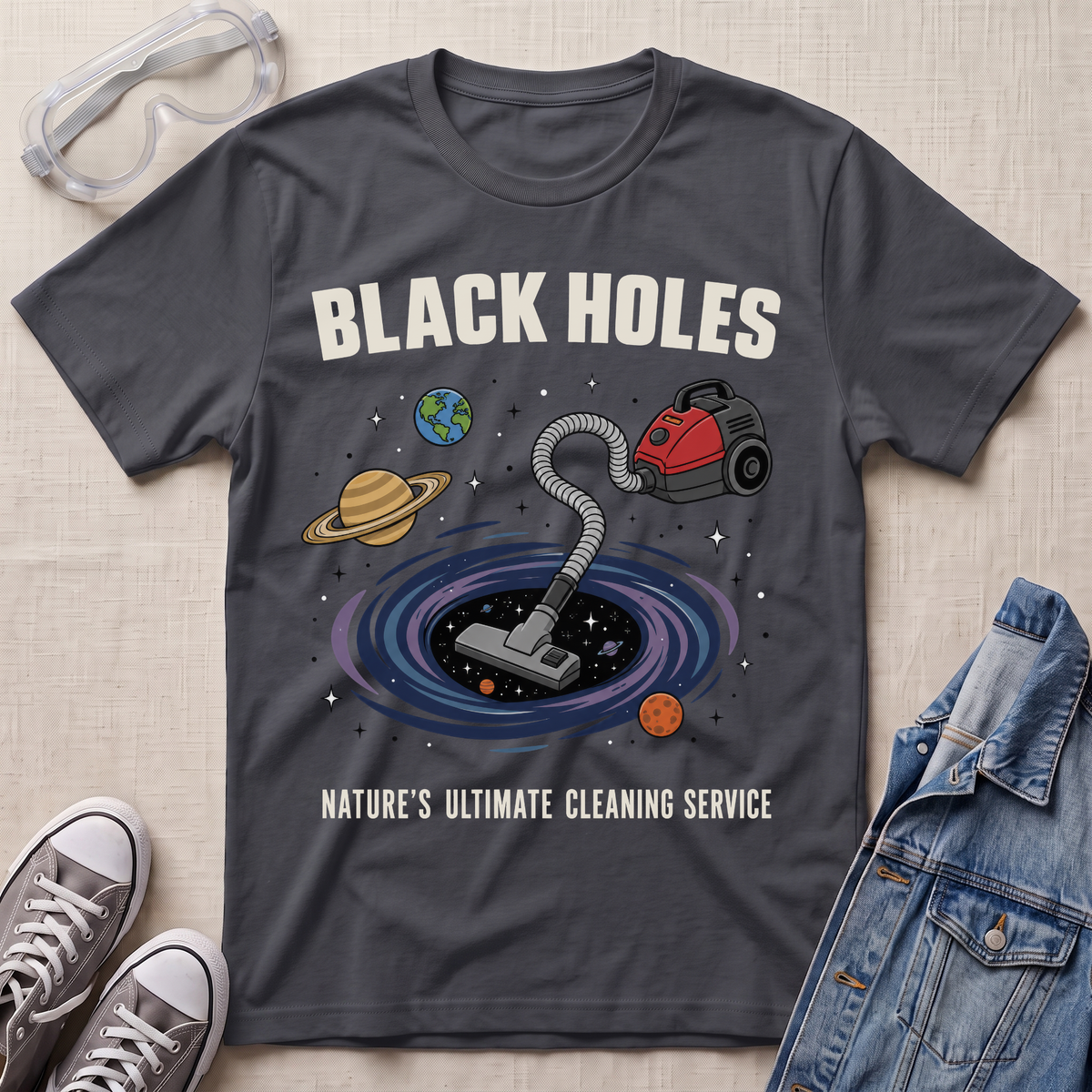 A charcoal graphic t-shirt laid flat on a textured fabric background, styled with safety goggles, sneakers, and a denim jacket. The t-shirt features a cartoon illustration of a vacuum cleaner falling into a black hole surrounded by planets, with the text 'BLACK HOLES' and 'NATURE'S ULTIMATE CLEANING SERVICE'.
