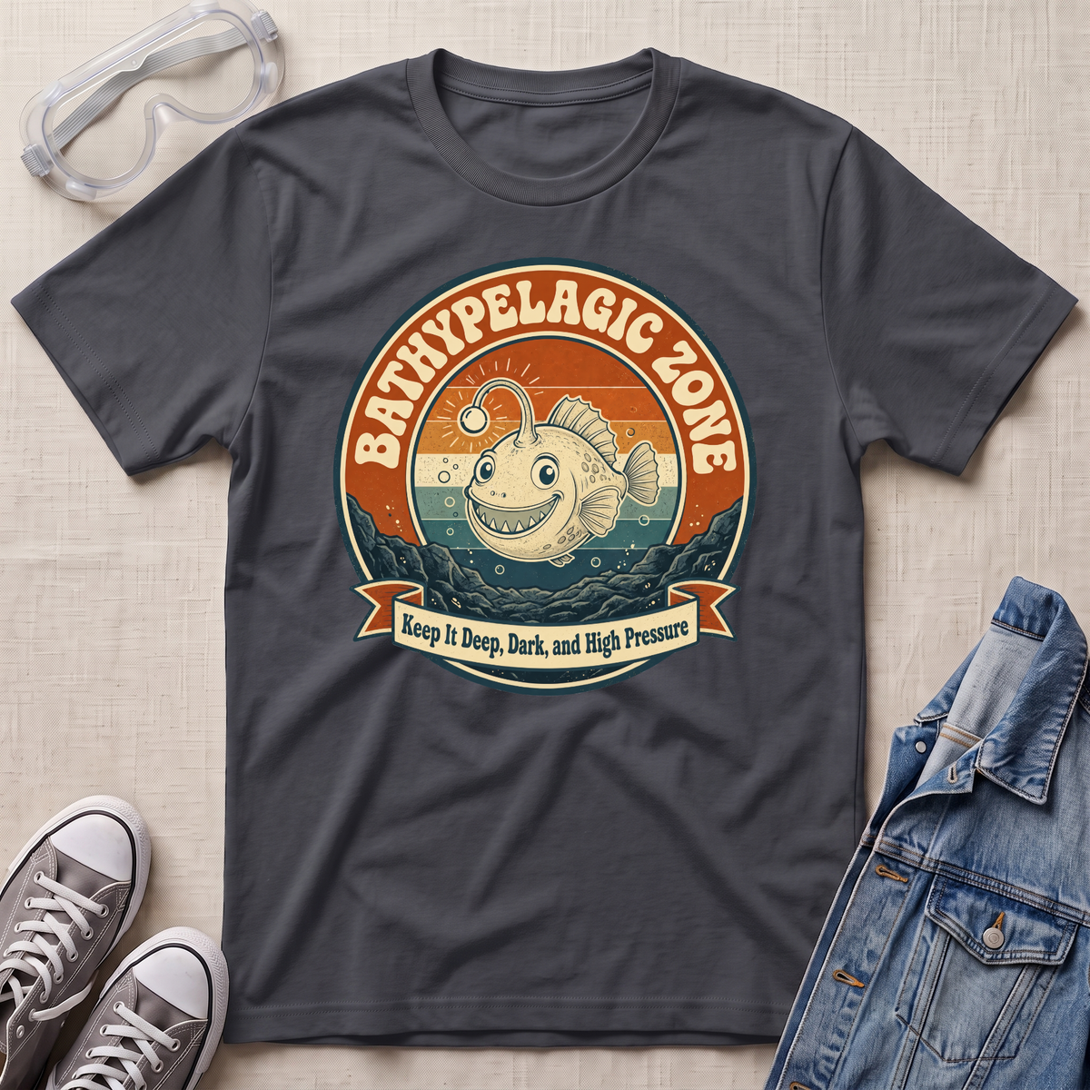 A dark charcoal gray graphic t-shirt laid flat on a textured beige fabric background, styled with safety goggles, gray sneakers, and a denim jacket. The t-shirt features a retro-style circular graphic of a smiling bathypelagic fish with the text 'BATHYPELAGIC ZONE' and 'Keep it Deep, Dark, and High Pressure'.