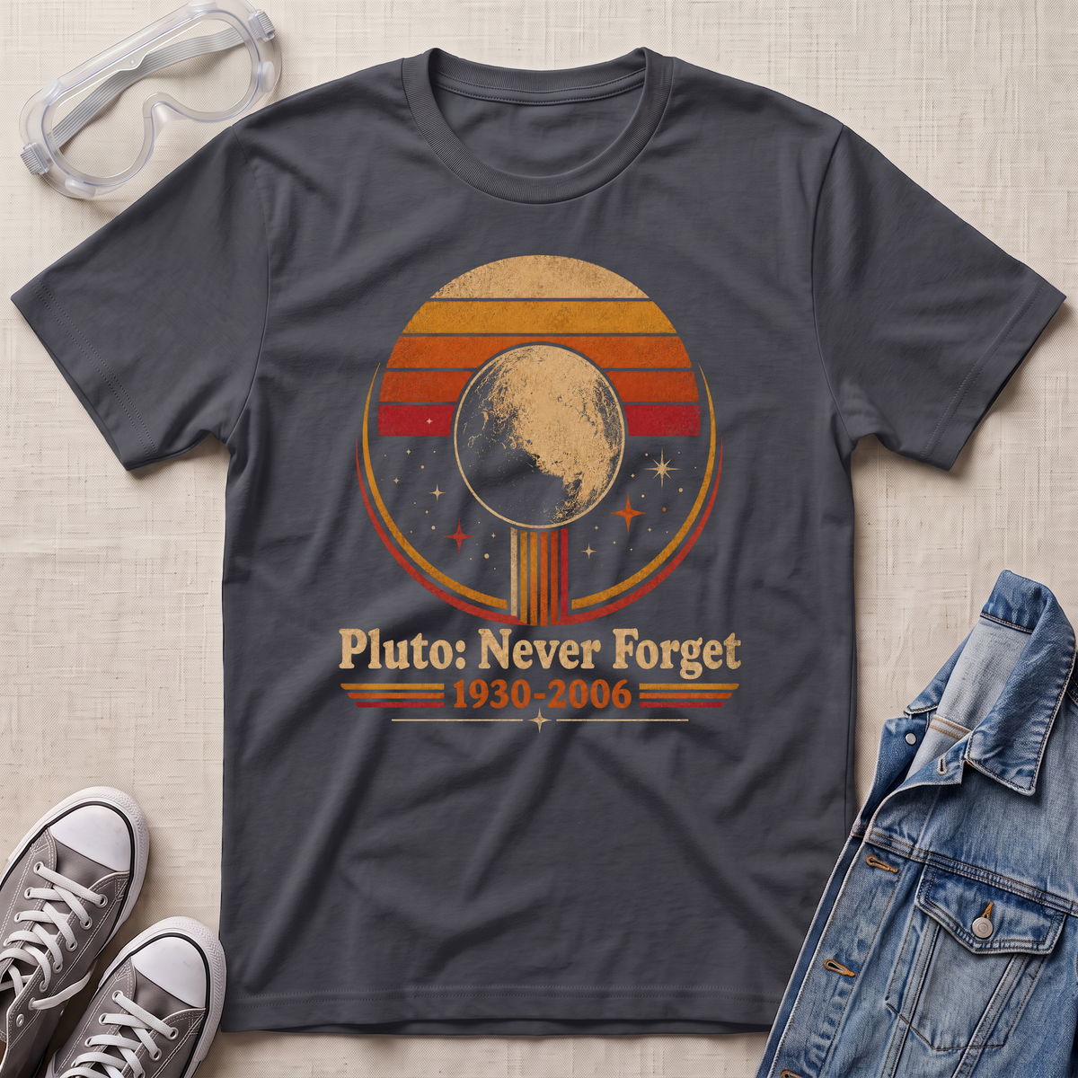 A dark charcoal gray graphic t-shirt laid flat on a textured beige fabric background, styled with clear safety goggles, gray sneakers, and a blue denim jacket. The t-shirt features a retro circular graphic of Pluto with the text 'Pluto: Never Forget 1930-2006'.