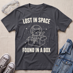 A charcoal gray graphic t-shirt laid flat on a beige fabric background, surrounded by styling props including clear safety glasses, gray sneakers, and a blue denim jacket. The t-shirt features a white graphic of a cat inside a box with a helmet and the text 'LOST IN SPACE FOUND IN A BOX'.