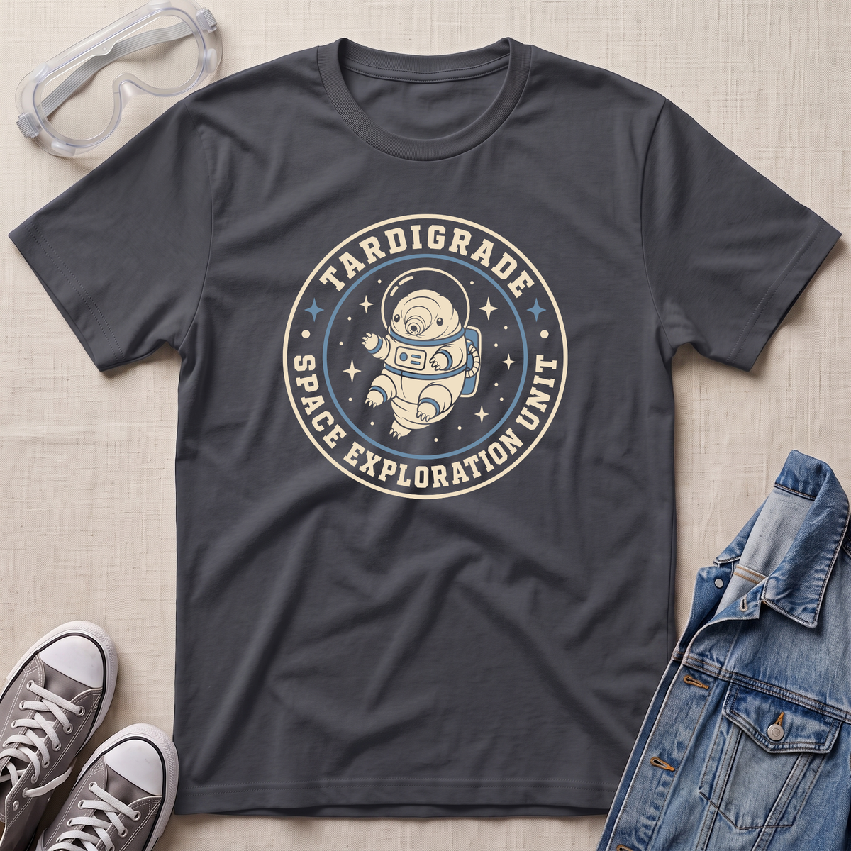 A dark charcoal gray graphic t-shirt featuring a retro circular astronaut design with the text 'TARDIGRADE SPACE EXPLORATION UNIT'. The t-shirt is styled in a flat-lay arrangement on a light beige textured background, accompanied by clear safety goggles, gray sneakers, and a blue denim jacket.