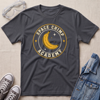 A dark charcoal gray t-shirt laid flat on a light beige textured background, featuring a circular graphic of a yellow banana in space with the words 'SPACE CHIMP ACADEMY'. The shirt is styled with props including clear safety goggles, gray sneakers, and a blue denim jacket.