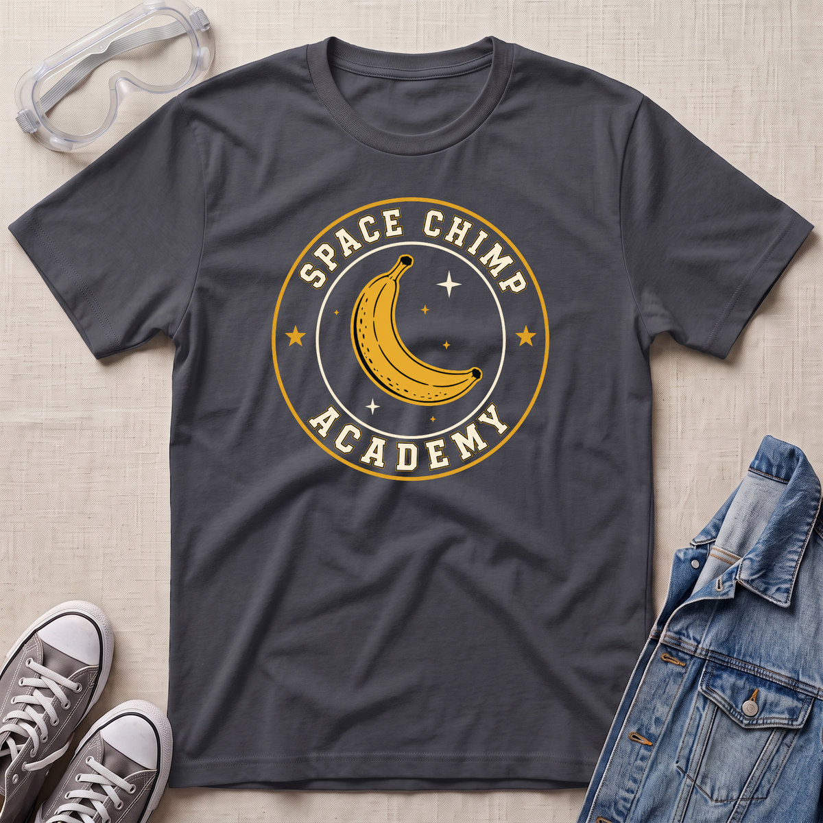 A dark charcoal gray t-shirt laid flat on a light beige textured background, featuring a circular graphic of a yellow banana in space with the words 'SPACE CHIMP ACADEMY'. The shirt is styled with props including clear safety goggles, gray sneakers, and a blue denim jacket.