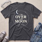 A charcoal gray graphic t-shirt laid flat on a textured beige fabric background, styled with clear safety goggles, gray sneakers, and a blue denim jacket. The t-shirt features a white circular graphic with a crescent moon and the text 'I'M OVER THE MOON AND STILL ORBITING'.