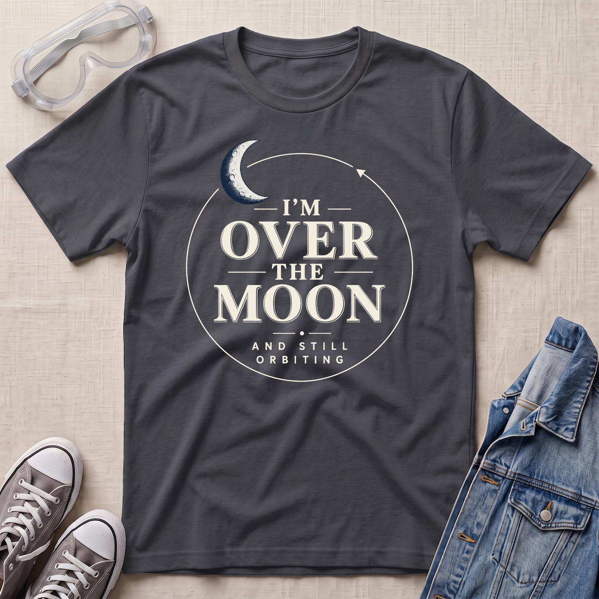 A charcoal gray graphic t-shirt laid flat on a textured beige fabric background, styled with clear safety goggles, gray sneakers, and a blue denim jacket. The t-shirt features a white circular graphic with a crescent moon and the text 'I'M OVER THE MOON AND STILL ORBITING'.