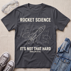 A charcoal gray graphic t-shirt featuring a white rocket science blueprint design with the text 'ROCKET SCIENCE IT'S NOT THAT HARD (Wait, yes it is.)' laid flat on a beige fabric background, styled with safety goggles, sneakers, and a denim jacket.