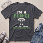 A dark charcoal gray graphic t-shirt laid flat on a light beige textured background, styled with safety goggles, gray sneakers, and a denim jacket. The t-shirt features a white and green graphic that reads 'I'M A FUN-GI BUT ONLY UNDER THE RIGHT CONDITIONS' with a mushroom illustration.