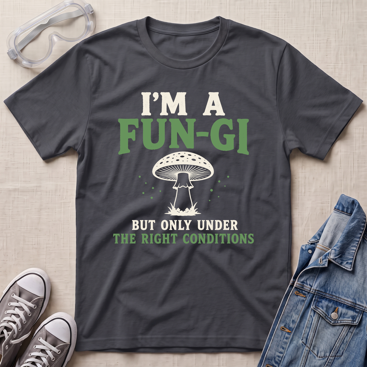 A dark charcoal gray graphic t-shirt laid flat on a light beige textured background, styled with safety goggles, gray sneakers, and a denim jacket. The t-shirt features a white and green graphic that reads 'I'M A FUN-GI BUT ONLY UNDER THE RIGHT CONDITIONS' with a mushroom illustration.