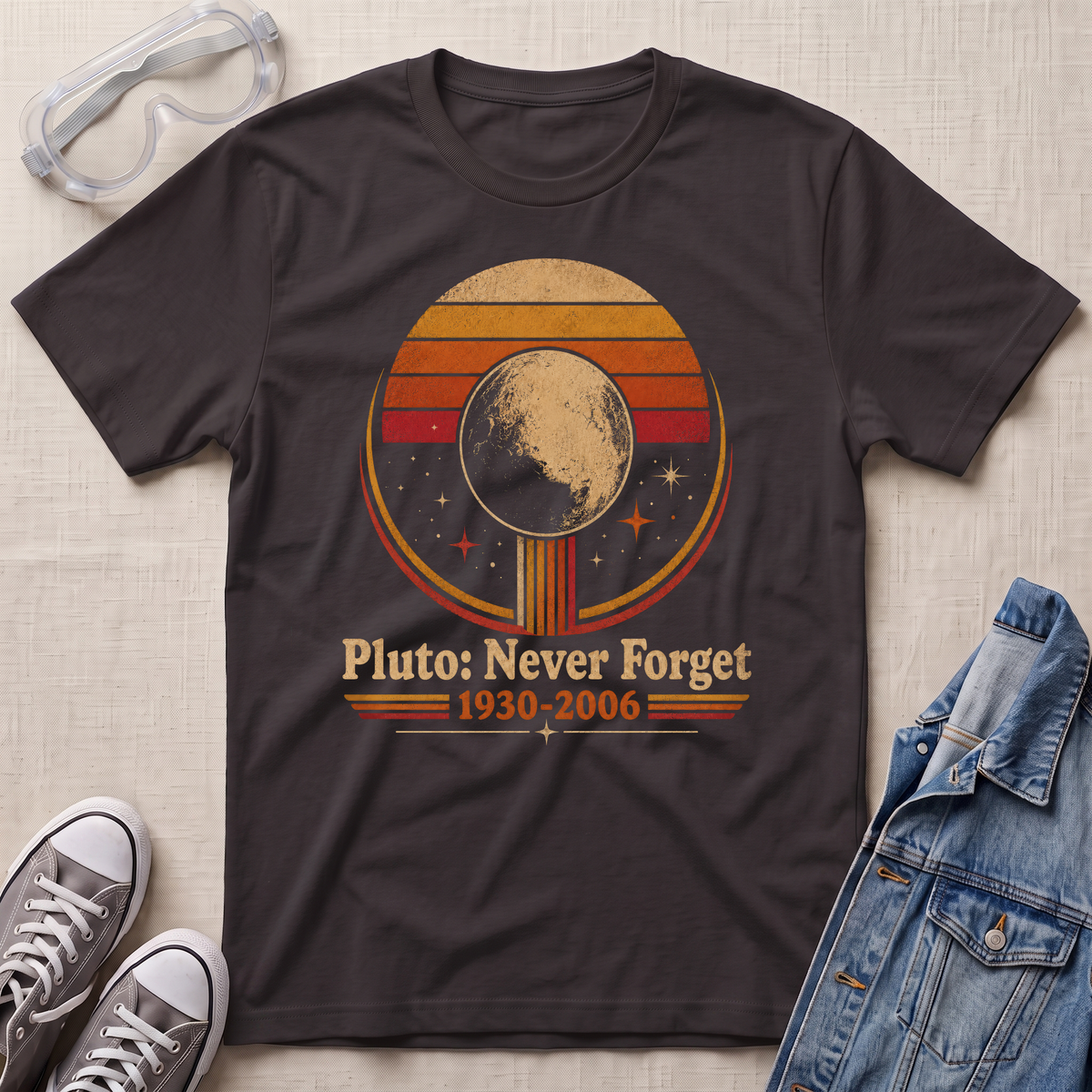 A dark chocolate brown graphic t-shirt laid flat on a light beige textured background, styled with safety goggles, gray sneakers, and a denim jacket. The t-shirt features a retro circular graphic of Pluto with the text 'Pluto: Never Forget 1930-2006'.