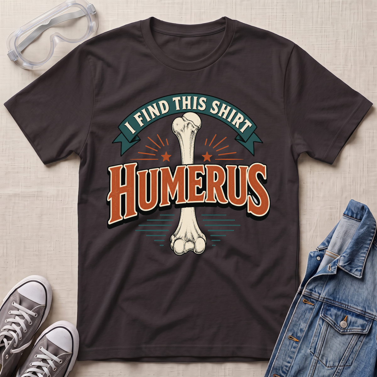 A dark chocolate brown graphic t-shirt laid flat on a light wood surface, featuring a bone graphic and the text 'I FIND THIS SHIRT HUMERUS' in orange and green. The shirt is styled with safety glasses, gray sneakers, and a denim jacket.
