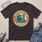 A dark chocolate brown graphic t-shirt laid flat on a light beige textured background, surrounded by lifestyle props including safety goggles, gray sneakers, and a denim jacket. The t-shirt features a circular retro-style graphic of a teal cartoon DNA molecule wearing a cap and holding a wrench, with the text 'DNA REPAIR SHOP' and 'Patching Your Base Pairs Since Day One'.