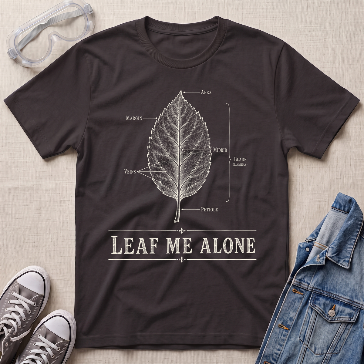 A dark chocolate brown botanical t-shirt featuring a vintage labeled leaf anatomy illustration with the pun “Leaf Me Alone” in bold retro typography.