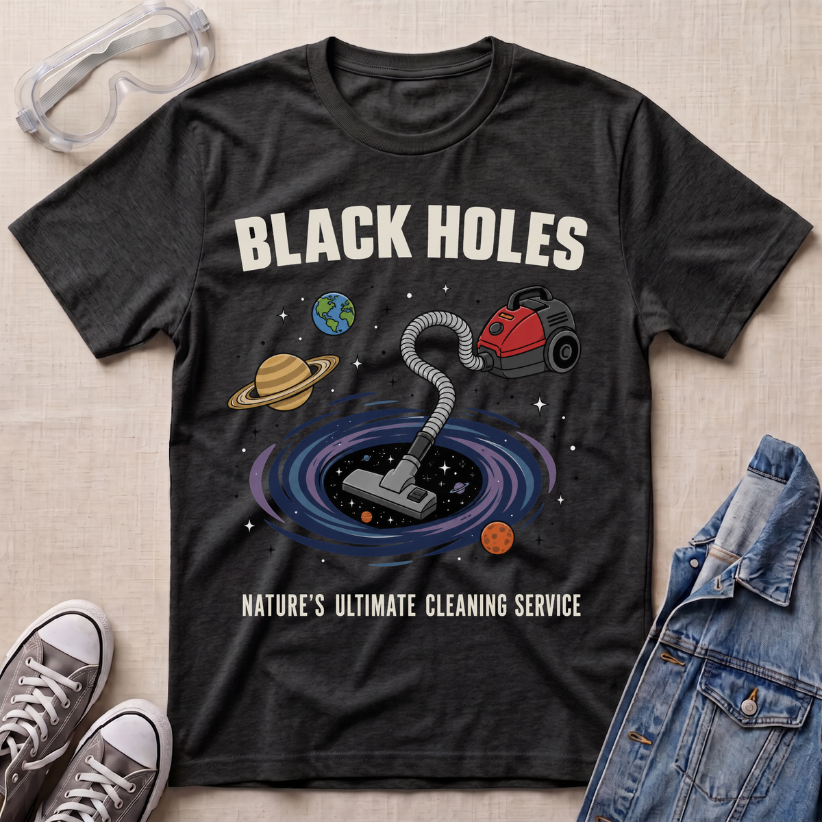 A dark heather grey graphic t-shirt laid flat on a textured fabric background, styled with safety goggles, sneakers, and a denim jacket. The t-shirt features a cartoon illustration of a vacuum cleaner falling into a black hole surrounded by planets, with the text 'BLACK HOLES' and 'NATURE'S ULTIMATE CLEANING SERVICE'.
