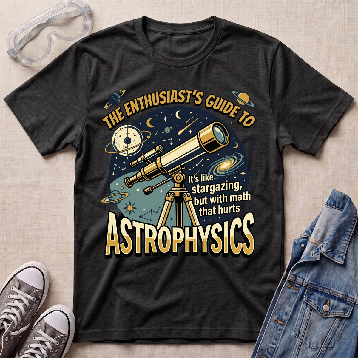 A dark heather graphic t-shirt laid flat on a beige fabric background, styled with safety glasses, gray sneakers, and a denim jacket. The t-shirt features a detailed space-themed graphic with a telescope, planets, stars, and text that reads 'THE ENTHUSIAST'S GUIDE TO ASTROPHYSICS'.