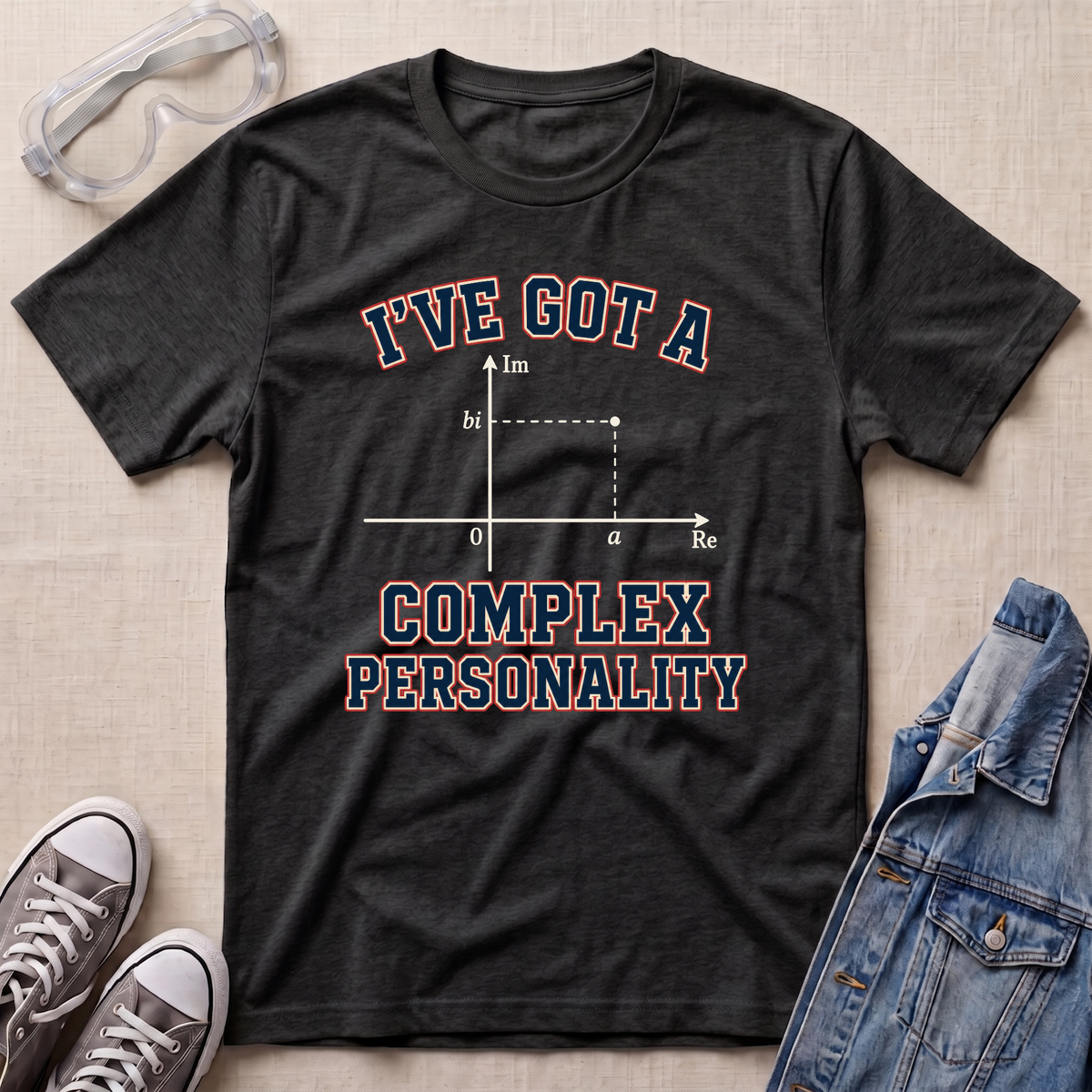 A dark heather gray graphic t-shirt laid flat on a beige fabric background, styled with safety goggles, gray sneakers, and a denim jacket. The t-shirt features a graphic of a complex impedance plot with the text 'I'VE GOT A COMPLEX PERSONALITY' in navy blue with a red outline.
