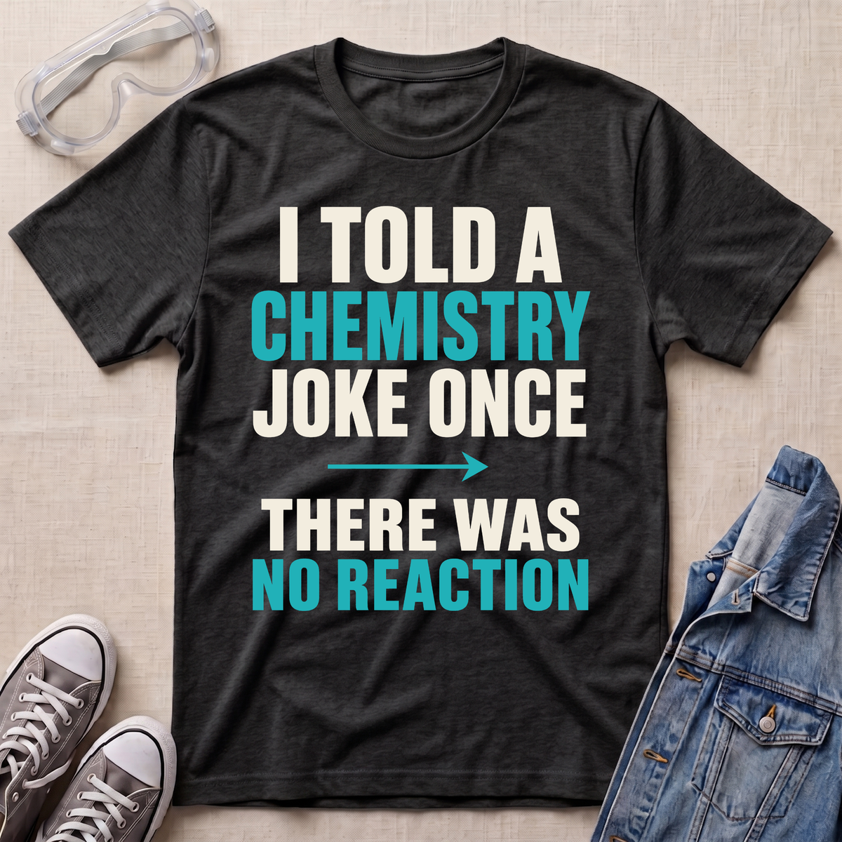 A dark heather gray graphic t-shirt laid flat on a beige textured background, surrounded by styling props including safety goggles, gray sneakers, and a denim jacket. The t-shirt features a bold text design in white and teal reading 'I TOLD A CHEMISTRY JOKE ONCE THERE WAS NO REACTION'.