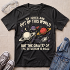 A dark heather gray graphic t-shirt laid flat on a beige textured background, styled with safety glasses, gray sneakers, and a denim jacket. The t-shirt features a humorous space-themed graphic with planets, stars, and the text 'MY JOKES ARE OUT OF THIS WORLD BUT THE GRAVITY OF THE SITUATION IS REAL'.