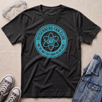 A dark heather gray graphic t-shirt laid flat on a beige textured background, styled with safety goggles, gray sneakers, and a denim jacket. The t-shirt features a teal circular graphic that reads 'HEISENBERG FAN CLUB' and 'WE'RE NOT CERTAIN WE'RE HERE.