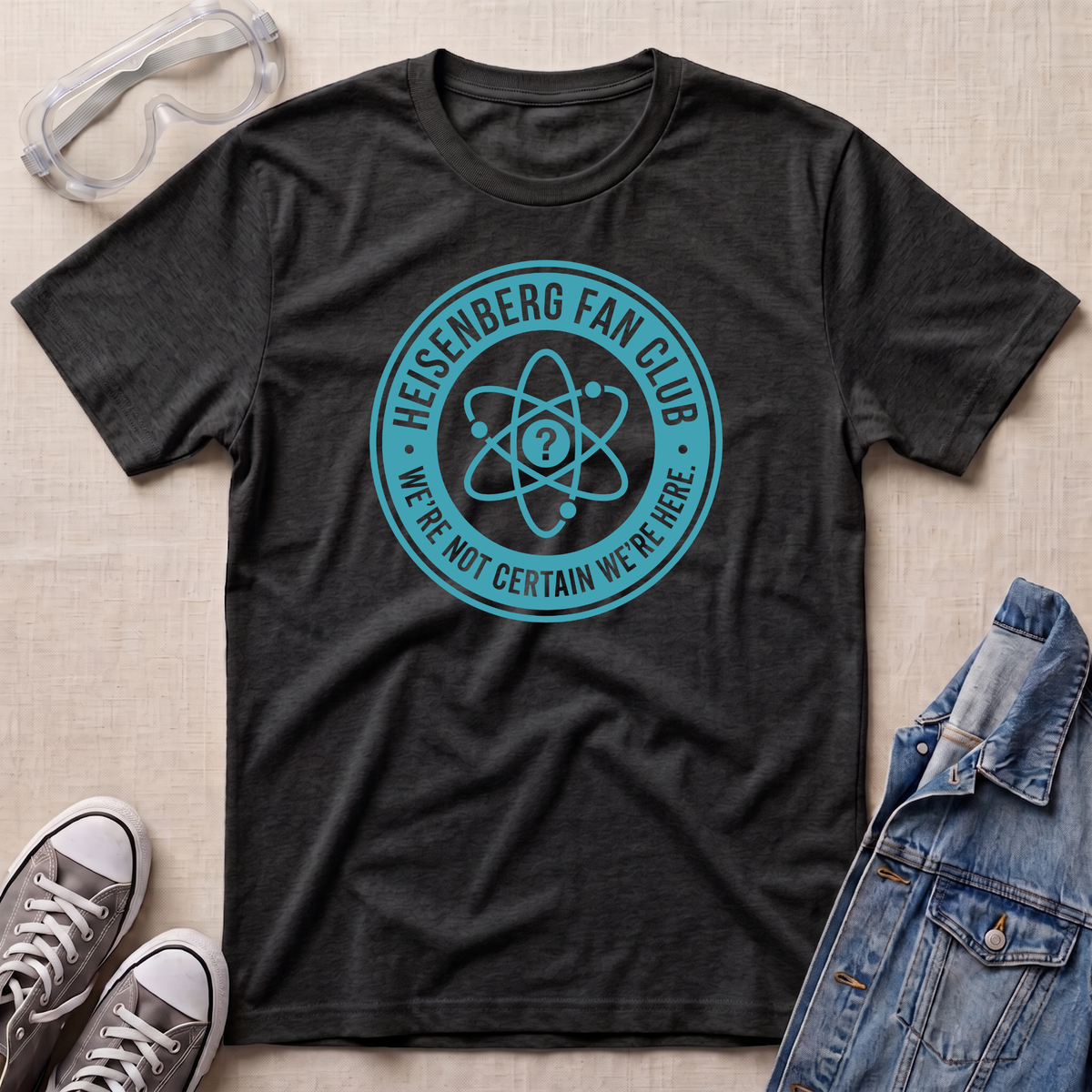 A dark heather gray graphic t-shirt laid flat on a beige textured background, styled with safety goggles, gray sneakers, and a denim jacket. The t-shirt features a teal circular graphic that reads 'HEISENBERG FAN CLUB' and 'WE'RE NOT CERTAIN WE'RE HERE.