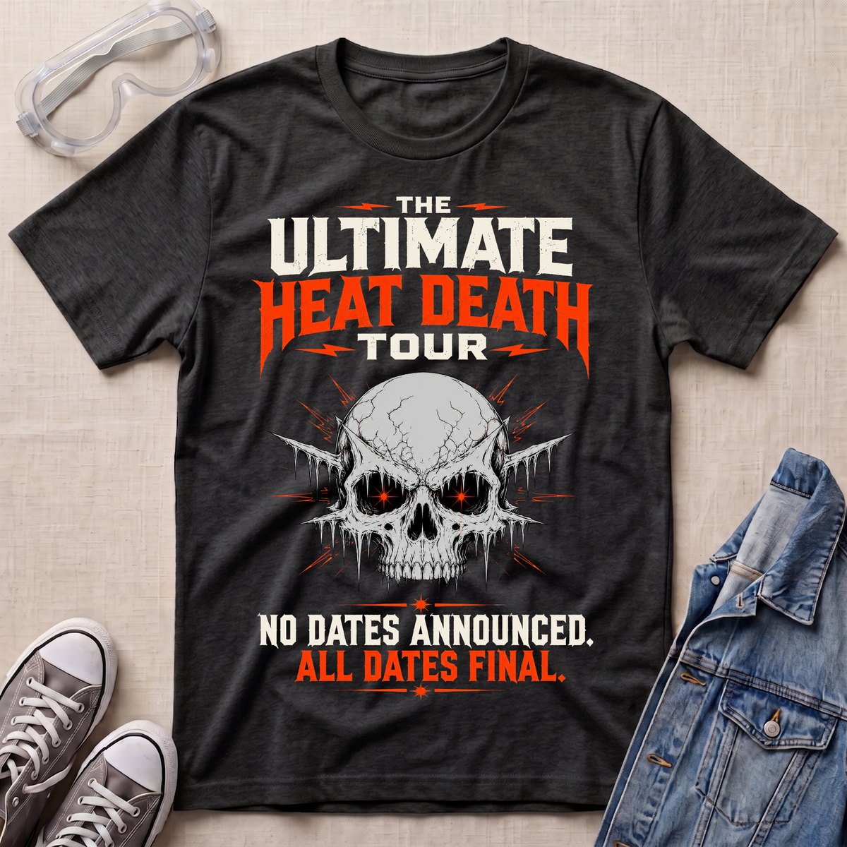 A dark heather gray graphic t-shirt laid flat on a beige fabric background, styled with safety glasses, gray sneakers, and a blue denim jacket. The t-shirt features a skull graphic with glowing red eyes and bold text that reads 'THE ULTIMATE HEAT DEATH TOUR' and 'NO DATES ANNOUNCED.