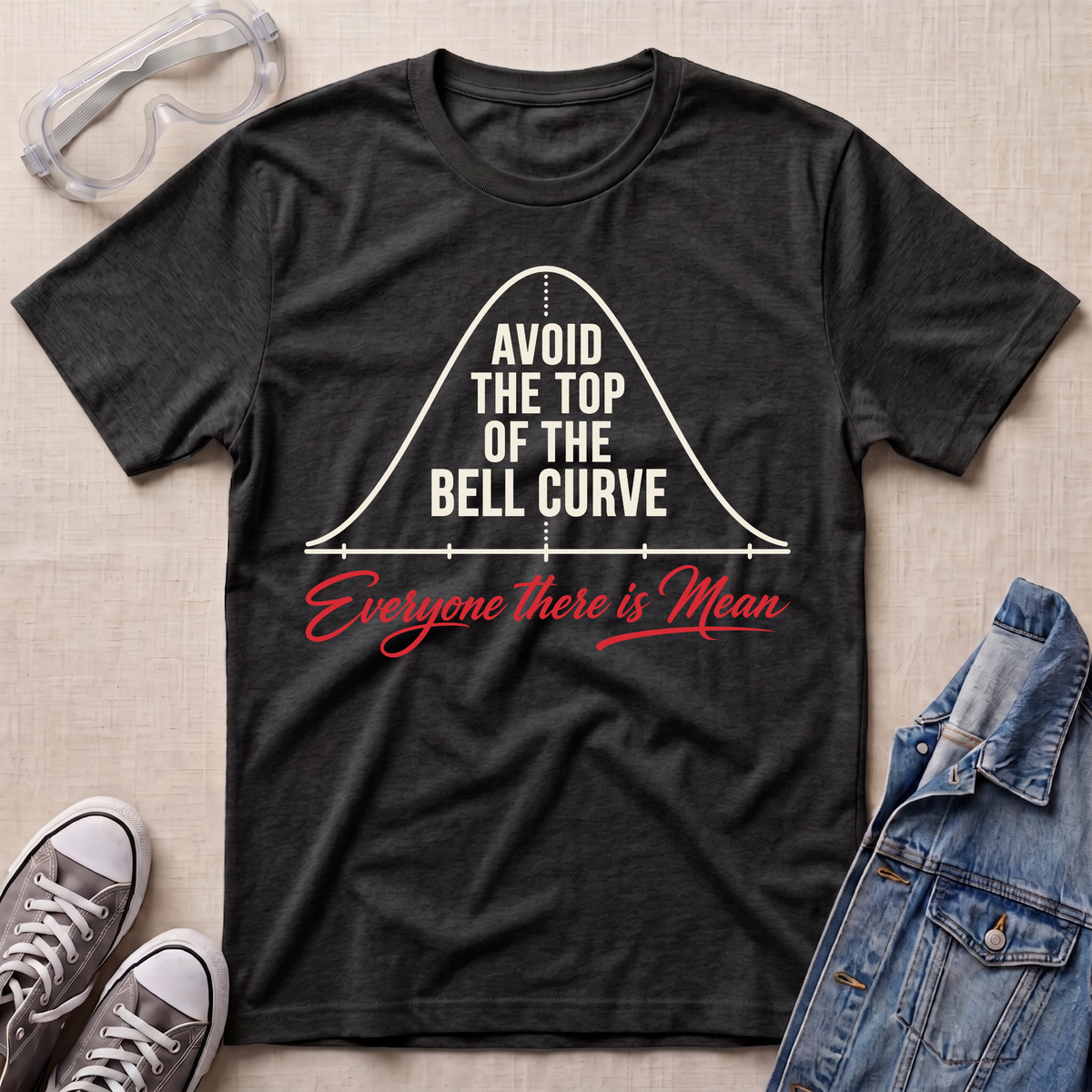 A dark heather gray graphic t-shirt laid flat on a beige fabric background, styled with safety glasses, gray sneakers, and a blue denim jacket. The t-shirt features a white bell curve graphic with the text 'AVOID THE TOP OF THE BELL CURVE' and red cursive text reading 'Everyone there is Mean'.