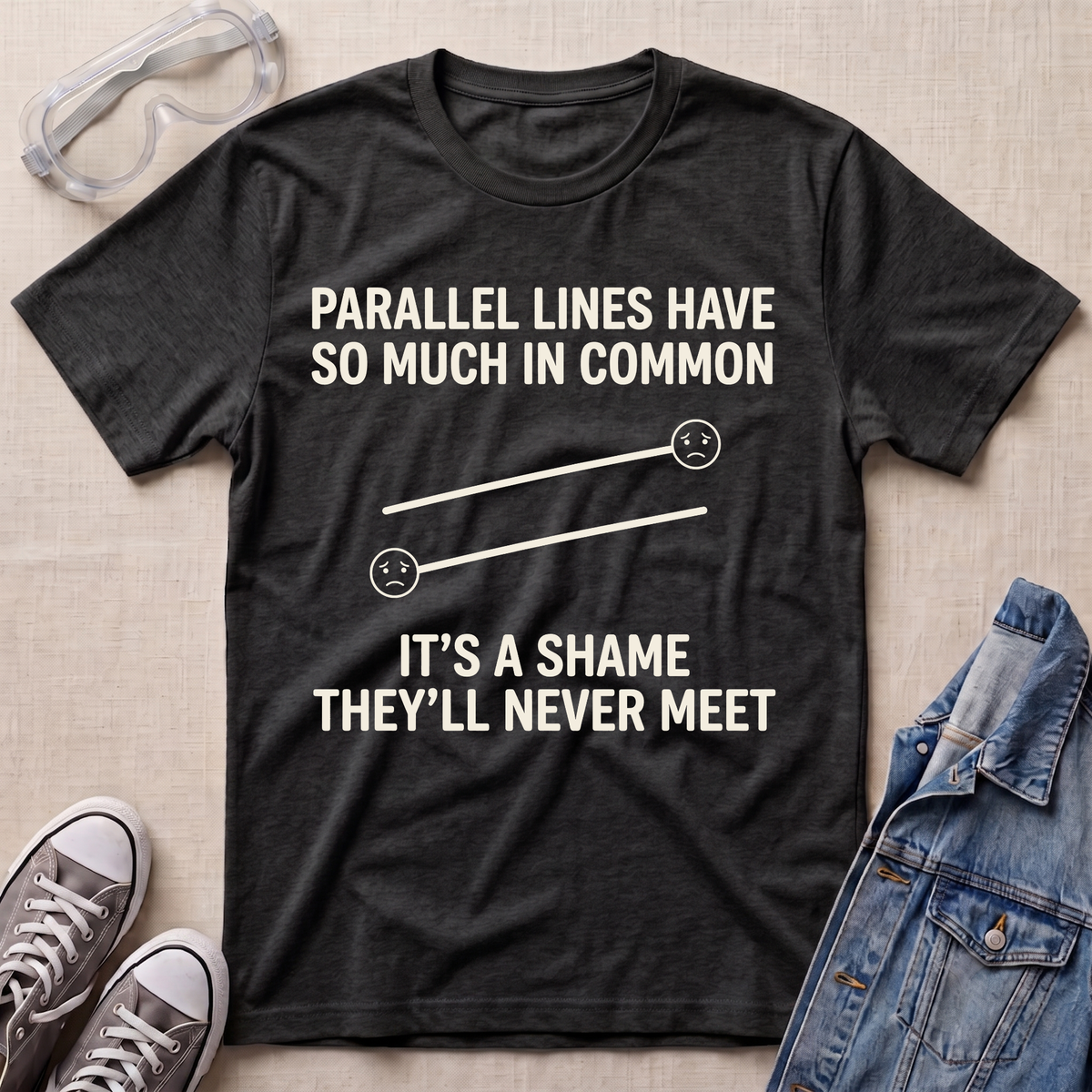 A dark heather gray graphic t-shirt laid flat on a beige fabric background, surrounded by styling props including safety goggles, gray sneakers, and a blue denim jacket. The t-shirt features a white printed graphic of two parallel sad faces with the text 'PARALLEL LINES HAVE SO MUCH IN COMMON' and 'IT'S A SHAME THEY'LL NEVER MEET'.