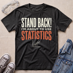 A dark charcoal heather graphic t-shirt laid flat on a beige textured background, surrounded by props including safety goggles, gray sneakers, and a denim jacket. The t-shirt features the text 'STAND BACK! I'M ABOUT TO USE STATISTICS' in white and orange lettering.