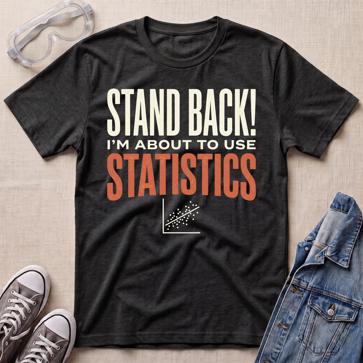 A dark charcoal heather graphic t-shirt laid flat on a beige textured background, surrounded by props including safety goggles, gray sneakers, and a denim jacket. The t-shirt features the text 'STAND BACK! I'M ABOUT TO USE STATISTICS' in white and orange lettering.
